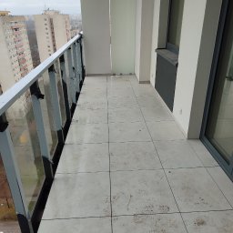 karmarRemonty Marcin Pański - Balkon w nowoczesnym apartamentowcu z wyłożoną jasnymi płytkami podłogą, przeszkloną balustradą i widokiem na inne budynki w tle, na płytkach widoczne ślady zabrudzeń.