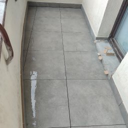 karmarRemonty Marcin Pański - Szare płytki ułożone na podłodze balkonu, widoczne ślady po fugowaniu, żółta gąbka na parapecie i kartonowe podkładki przy oknie.