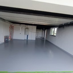 karmarRemonty Marcin Pański - Wnętrze w trakcie prac wykończeniowych z świeżo wylaną, szarą posadzką żywiczną, zabezpieczoną taśmą malarską przy ścianach, widoczne instalacje na ścianie i surowy stan wykończenia.