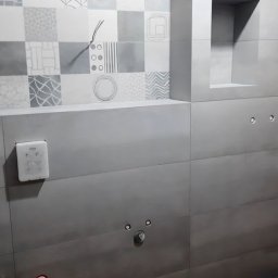 karmarRemonty Marcin Pański - Ściana łazienki w trakcie wykańczania z patchworkiem płytek w odcieniach szarości i bieli, widoczne wystające przewody, otwory na armaturę i fragment drewnianej podłogi.