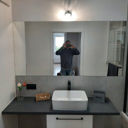 karmarRemonty Marcin Pański - Nowoczesna łazienka z dużym lustrem, w którym odbija się osoba robiąca zdjęcie telefonem; na blacie umywalka nablatowa, dekoracje i pojemnik na akcesoria łazienkowe.