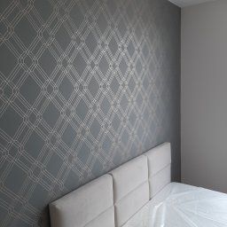 karmarRemonty Marcin Pański - Pokój z szarą tapetą w geometryczne wzory i łóżkiem z białym zagłówkiem, materac zabezpieczony folią ochronną.