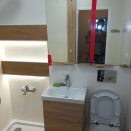 karmarRemonty Marcin Pański - Nowoczesna łazienka w trakcie montażu, widoczna umywalka z szafką w kolorze drewna, biała toaleta, brodzik i lustro zabezpieczone czerwoną taśmą malarską, odbicie osoby robiącej zdjęcie.