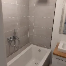 Wykończona łazienka z beżowymi, poziomymi płytkami, białą wanną i chromowaną baterią prysznicową; widoczna umywalka z drewnianym blatem.