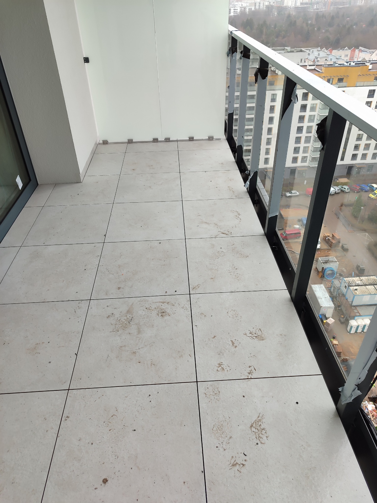 Balkon z jasnymi płytkami podłogowymi, śladami brudu i częściowo odklejoną folią ochronną na szklanej balustradzie, z widokiem na zabudowę miejską w tle.
