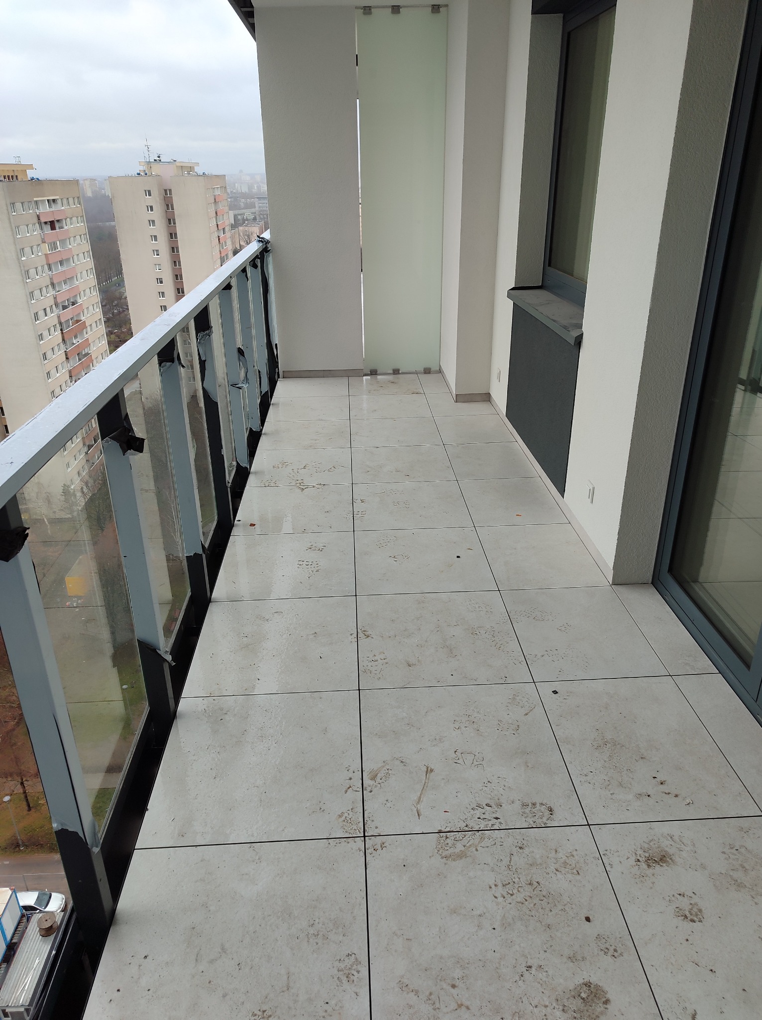 Balkon w nowoczesnym apartamentowcu z wyłożoną jasnymi płytkami podłogą, przeszkloną balustradą i widokiem na inne budynki w tle, na płytkach widoczne ślady zabrudzeń.