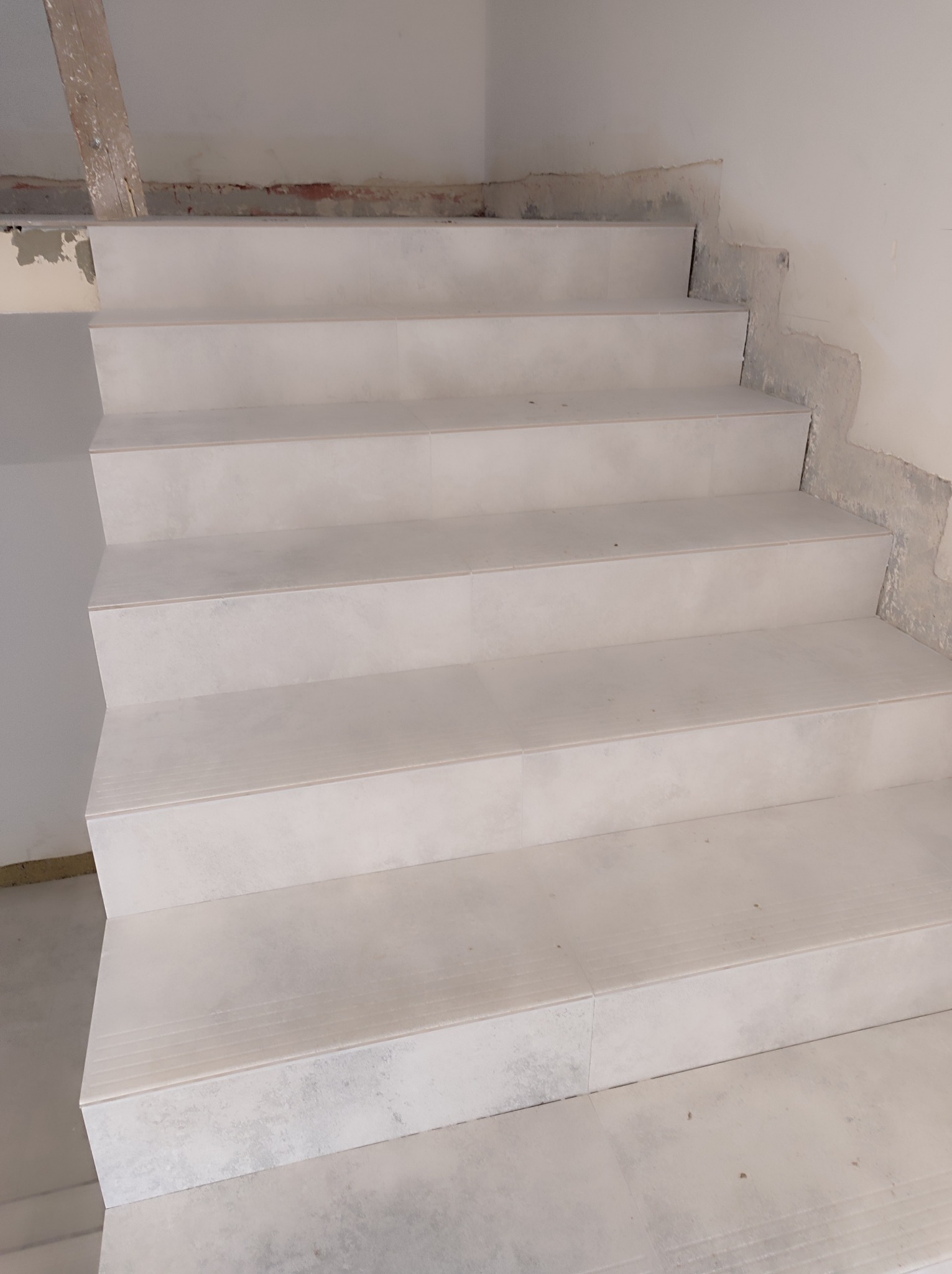 Schody wyłożone jasnymi, matowymi płytkami imitującymi kamień; widoczne niedokończone wykończenia ścian i sufitu.