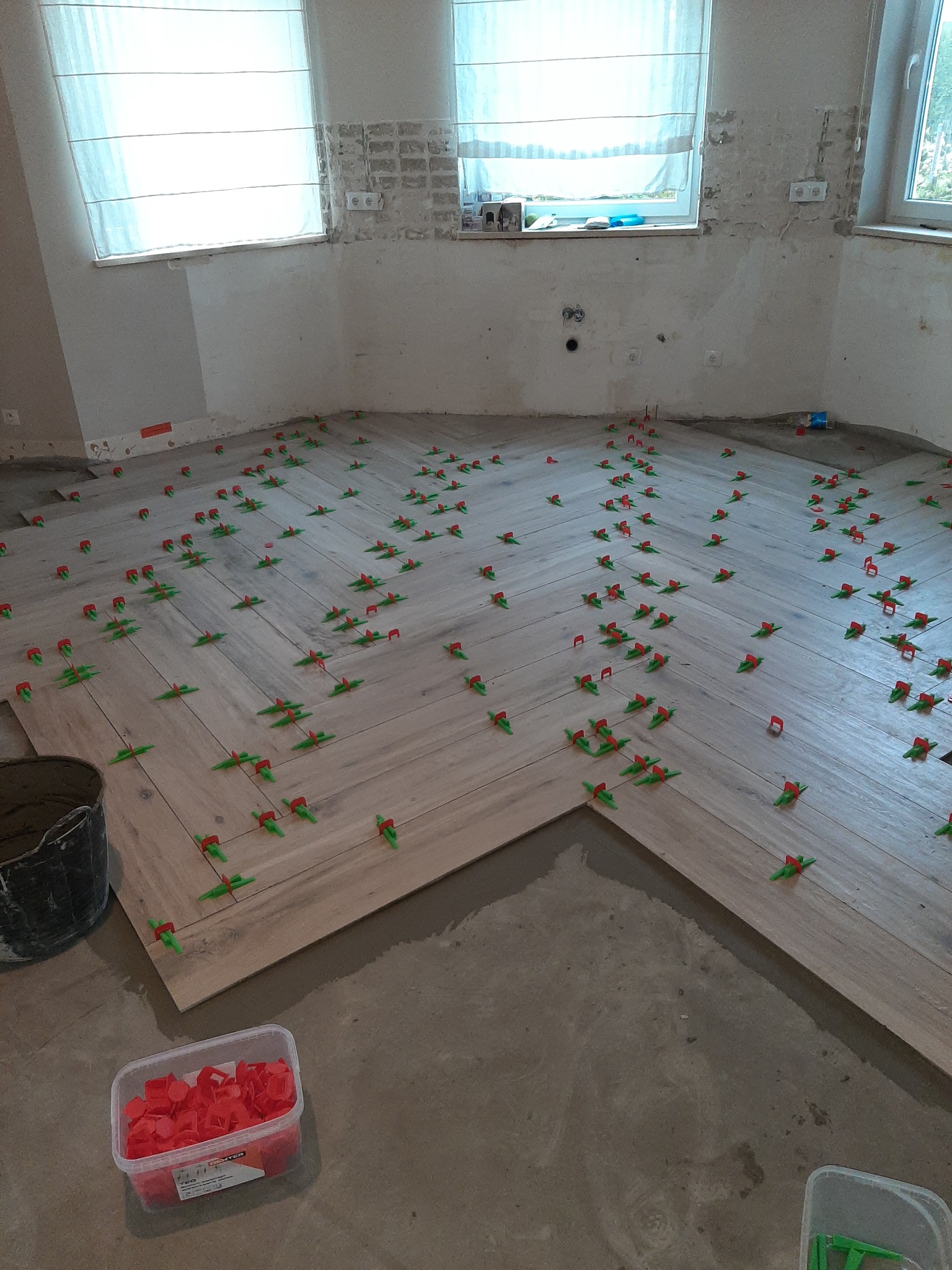 Układanie podłogi z płytek imitujących drewno w jodełkę, z użyciem czerwono-zielonych krzyżyków dystansowych, widoczne okna z białymi zasłonami i surowe ściany w trakcie remontu.