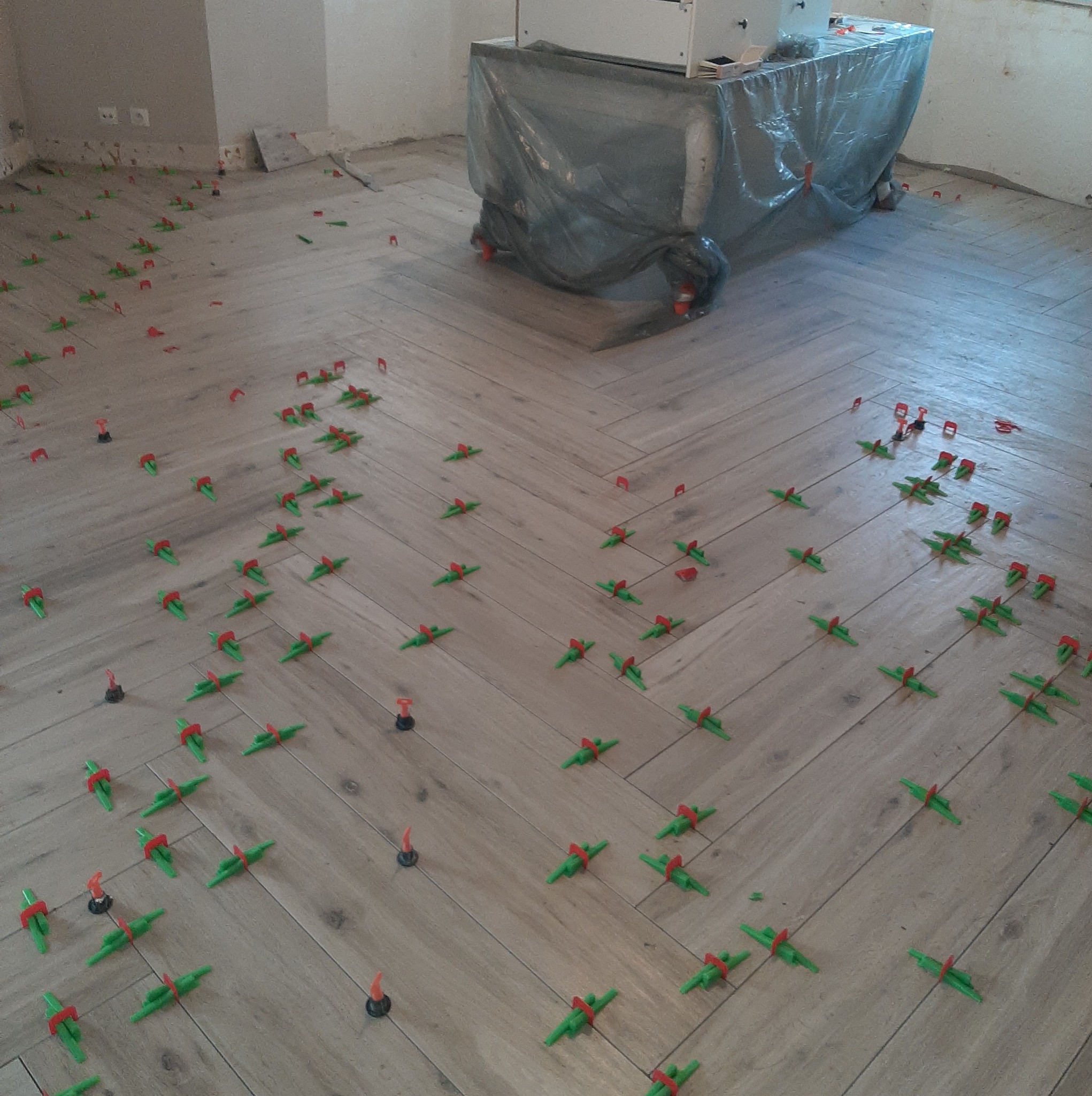 Ułożona pod kątem jasna podłoga z płytek imitujących drewno, z systemem poziomowania płytek (czerwono-zielone klipsy) na spoinach, w tle zakryty folią mebel.