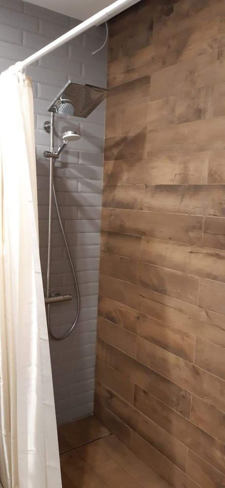 Nowoczesna kabina prysznicowa z szarą, cegiełkową glazurą i panelami imitującymi drewno, zasłonięta częściowo białą zasłoną, widoczny duży deszczownica i bateria.