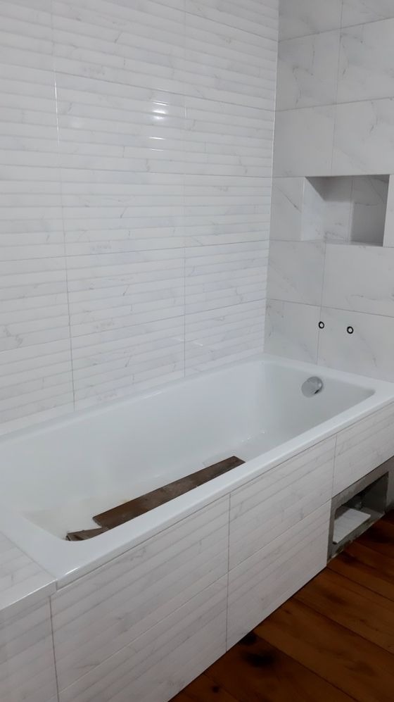 Biała wanna obudowana płytkami imitującymi marmur, z wnęką w ścianie i fragmentem drewnianej podłogi, w trakcie prac wykończeniowych.