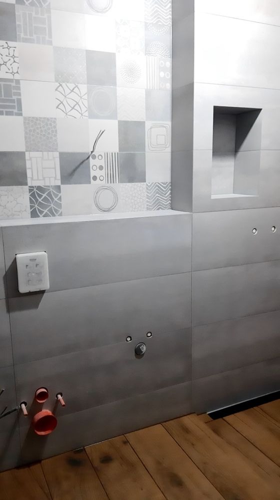 Ściana łazienki w trakcie wykańczania z patchworkiem płytek w odcieniach szarości i bieli, widoczne wystające przewody, otwory na armaturę i fragment drewnianej podłogi.