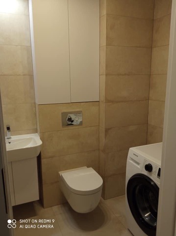 Nowoczesna łazienka z beżowymi płytkami, wiszącą miską WC, umywalką z szafką i pralką Samsung.