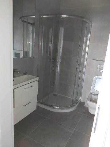 Nowoczesna łazienka z szarą glazurą podłogową, białą szafką pod umywalką i kabiną prysznicową z zaokrąglonymi szybami.