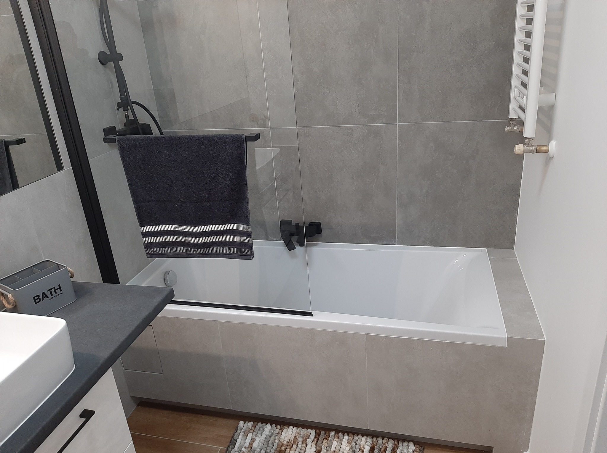 Nowoczesna łazienka z wanną obudowaną szarą płytką, czarną armaturą i grzejnikiem drabinkowym. W tle widoczna ściana wyłożona dużymi, szarymi płytkami.