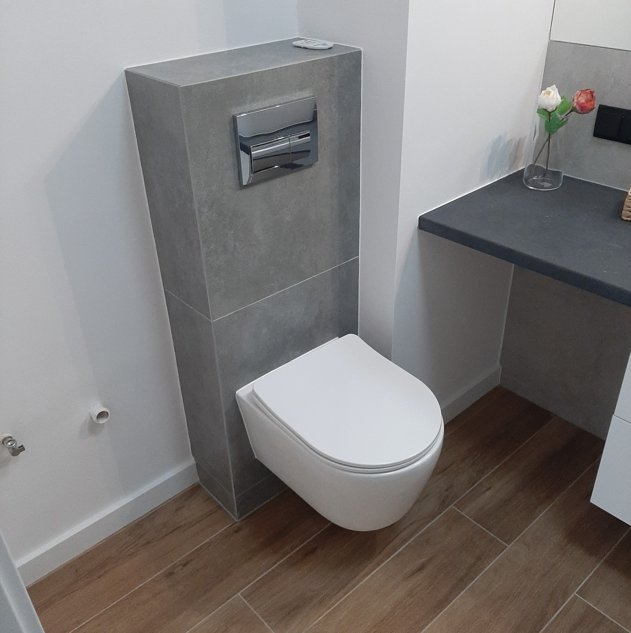 Nowoczesna, podwieszana toaleta z szarym, kamiennym stelażem i chromowanym przyciskiem spłukującym, drewnopodobne płytki podłogowe, minimalistyczna umywalka z kwiatami w tle.
