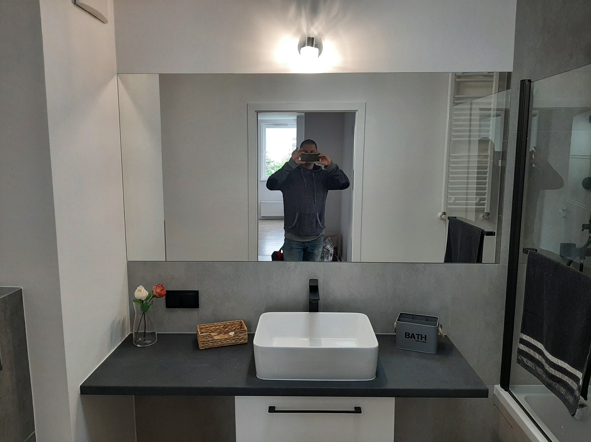 Nowoczesna łazienka z dużym lustrem, w którym odbija się osoba robiąca zdjęcie telefonem; na blacie umywalka nablatowa, dekoracje i pojemnik na akcesoria łazienkowe.