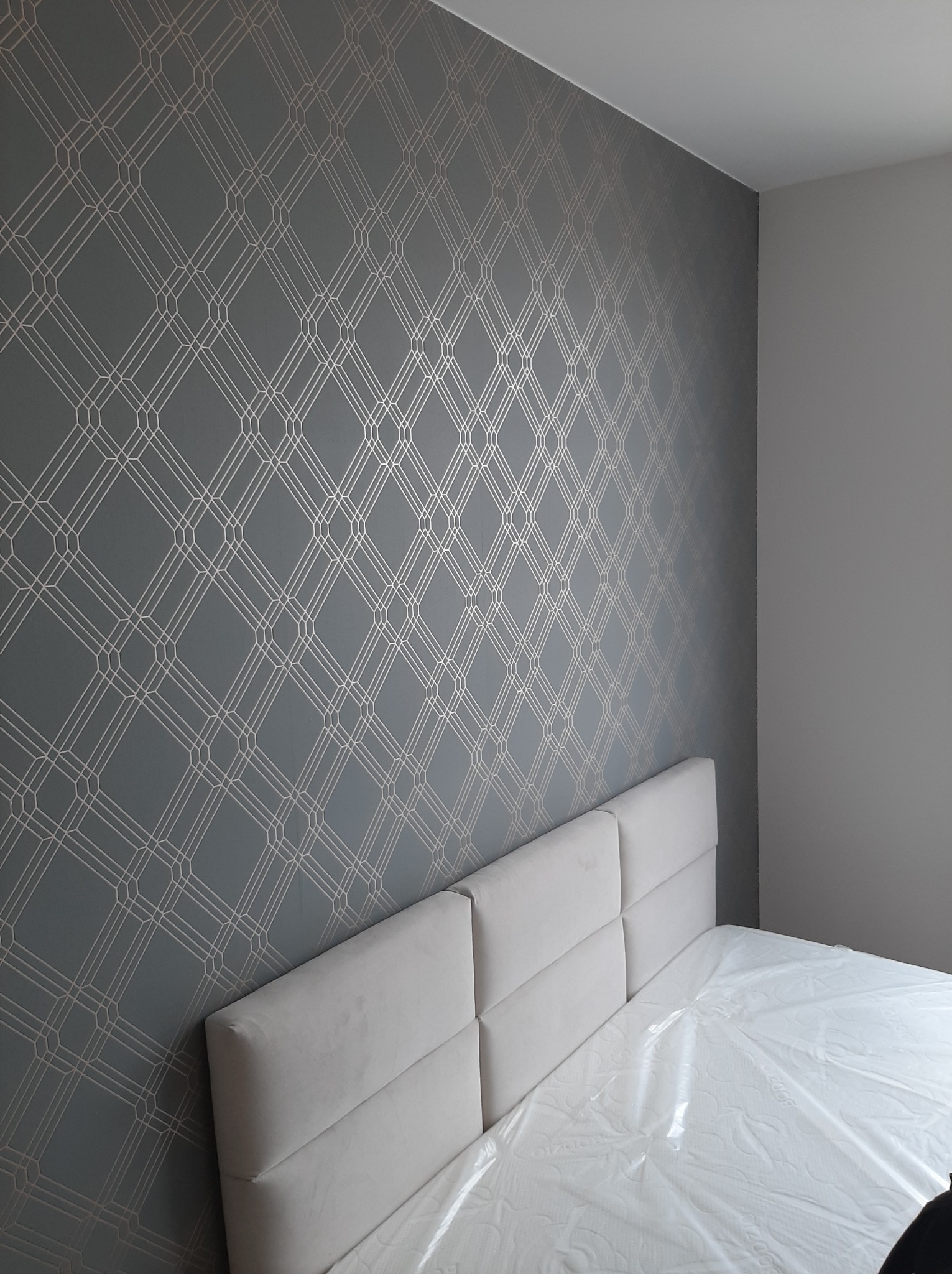 Pokój z szarą tapetą w geometryczne wzory i łóżkiem z białym zagłówkiem, materac zabezpieczony folią ochronną.