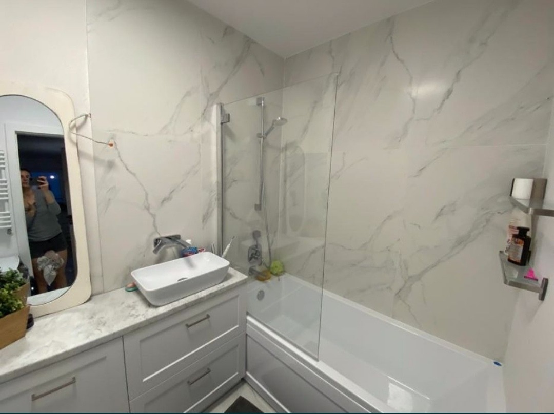 Nowoczesna łazienka z wanną za szklanym parawanem, umywalką nablatową i dużymi płytkami imitującymi marmur na ścianach, odbicie osoby robiącej zdjęcie w lustrze.