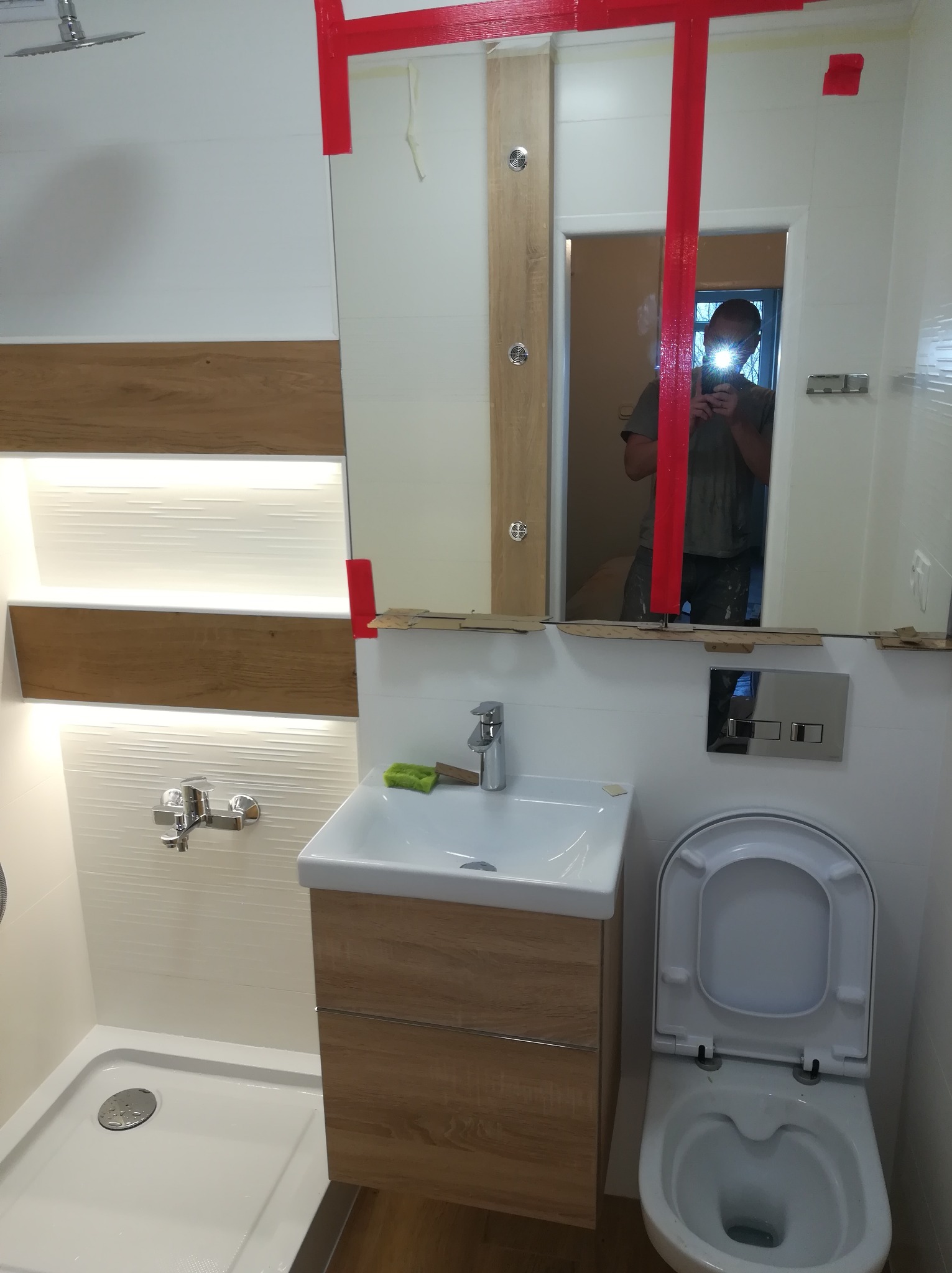 Nowoczesna łazienka w trakcie montażu, widoczna umywalka z szafką w kolorze drewna, biała toaleta, brodzik i lustro zabezpieczone czerwoną taśmą malarską, odbicie osoby robiącej zdjęcie.