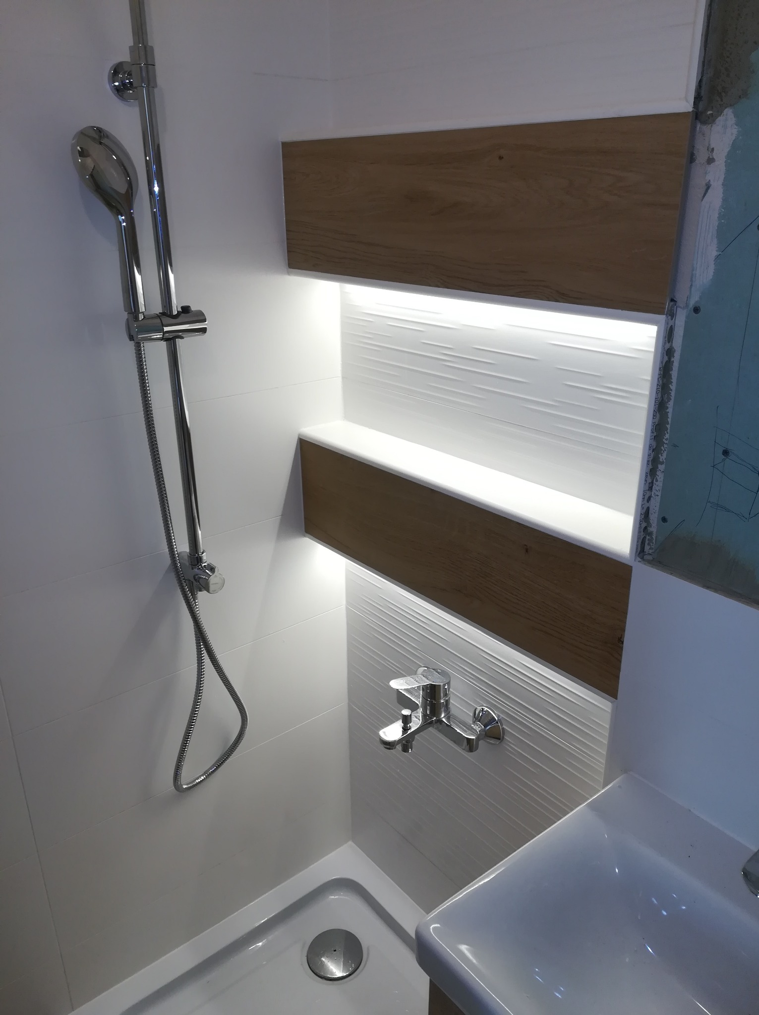 Nowoczesna kabina prysznicowa z drewnianymi półkami i oświetleniem LED w Warszawie, widoczna instalacja wodna i fragment umywalki.