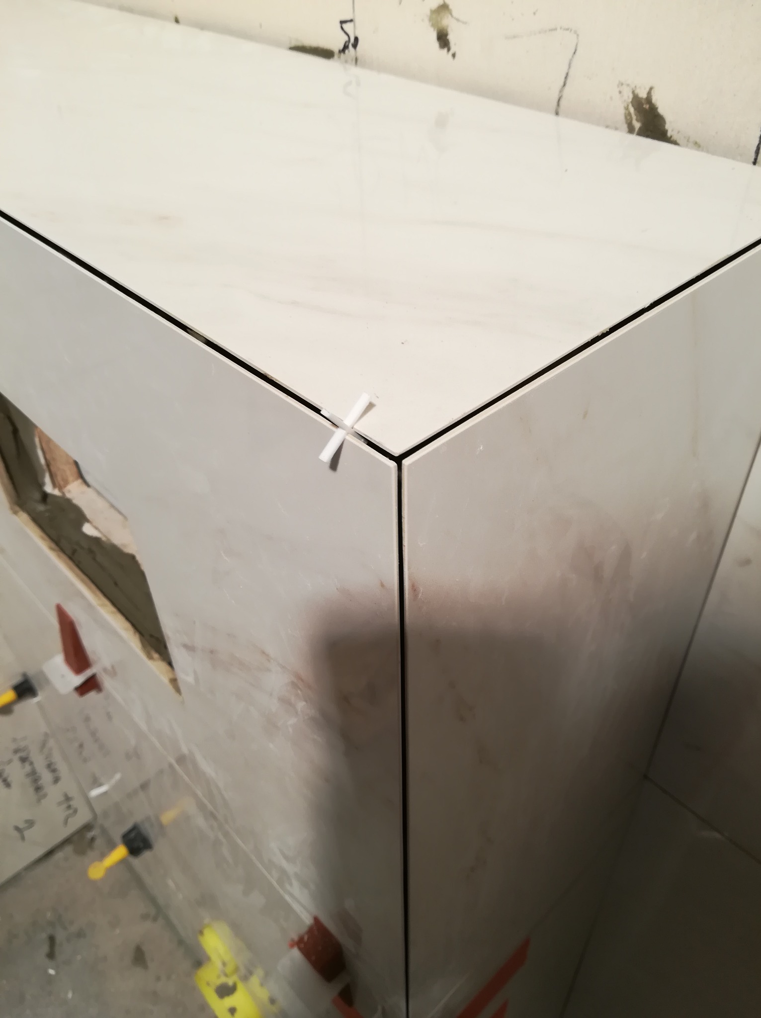 Fragment obudowy wanny lub umywalki z białych płytek imitujących marmur, z krzyżykiem dystansowym w rogu i widocznym otworem na instalację.