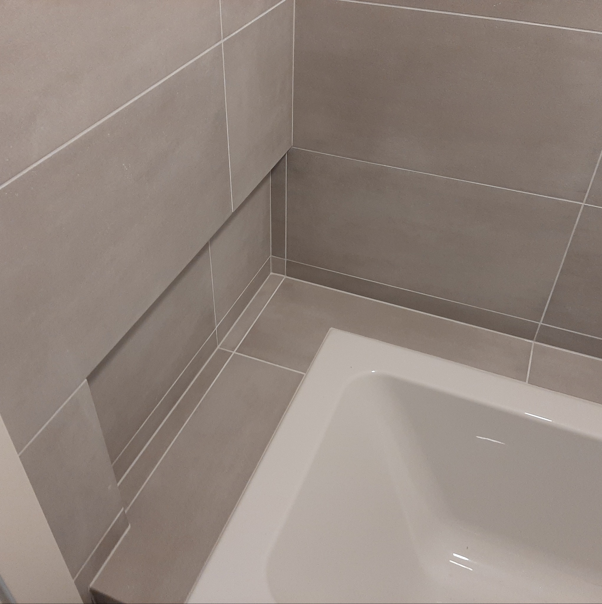 Fragment wykończonej łazienki z beżowymi płytkami i białą wanną, widok z góry na narożnik wanny i ściany.