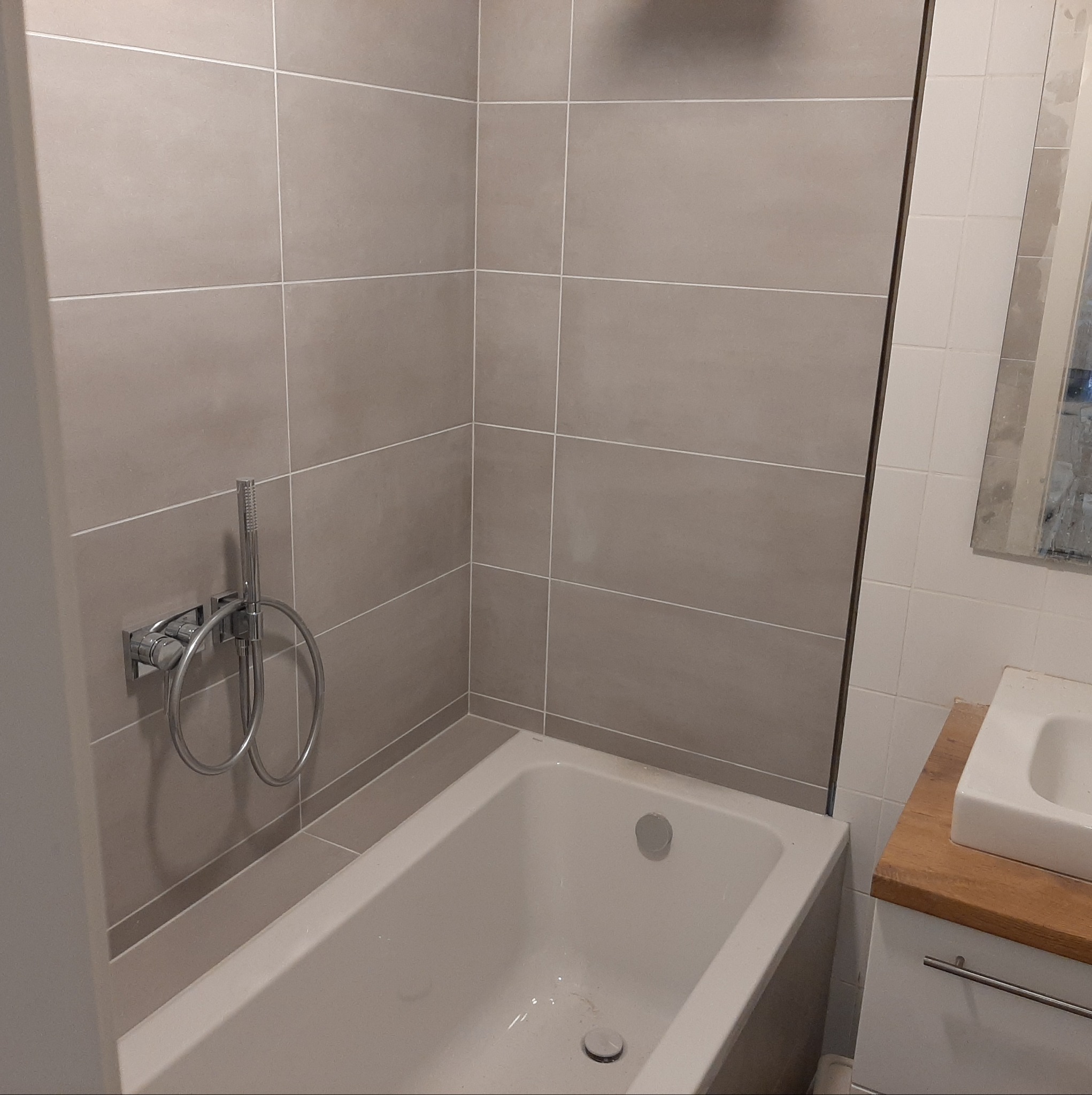 Wykończona łazienka z beżowymi, poziomymi płytkami, białą wanną i chromowaną baterią prysznicową; widoczna umywalka z drewnianym blatem.