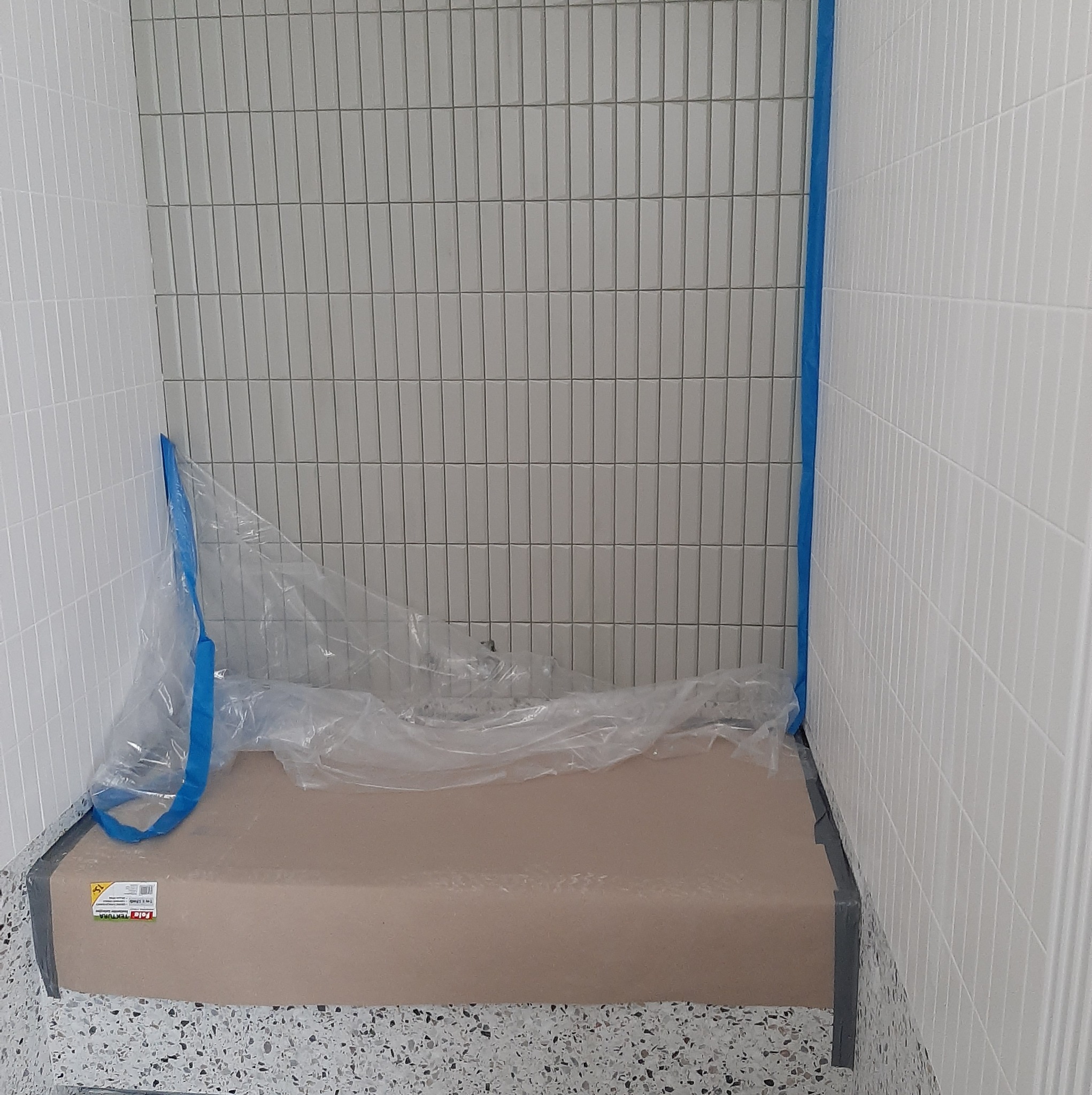 Przygotowanie do fugowania: Ściany w łazience wyłożone białymi i szarymi, wąskimi płytkami, podłoga zabezpieczona kartonem i folią ochronną, krawędzie zabezpieczone niebieską taśmą malarską.