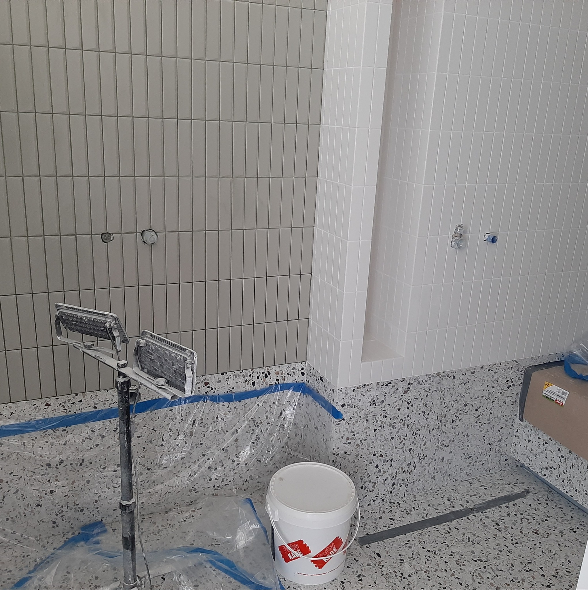 Wnętrze łazienki w trakcie prac wykończeniowych, widoczne pionowo ułożone, szare i białe płytki na ścianach, narzędzia glazurnicze i wiadro z farbą na zabezpieczonej folią podłodze.