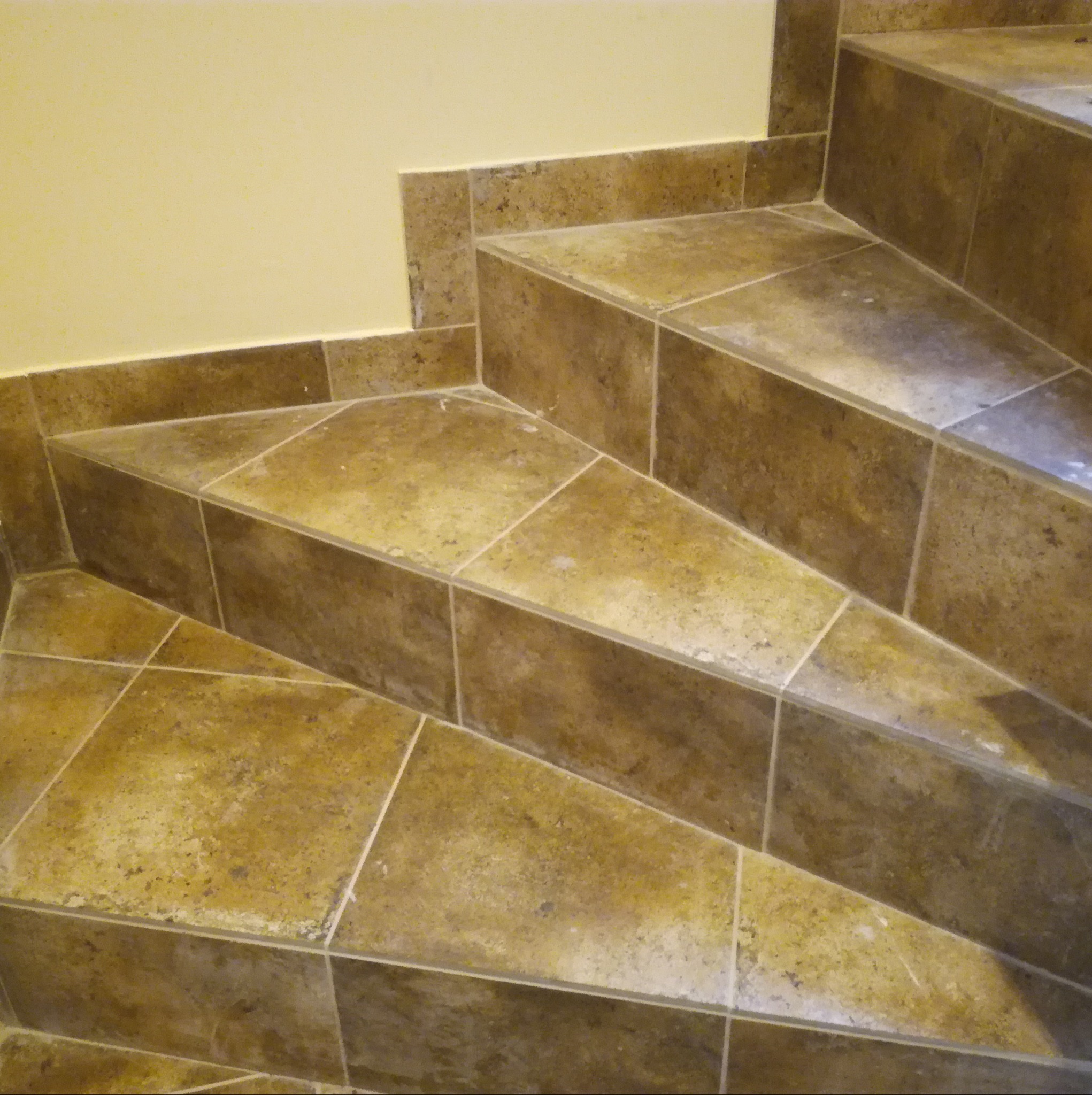 Schody wewnętrzne wyłożone brązowymi, kwadratowymi płytkami ceramicznymi z jasnymi fugami, przylegające do żółtej ściany.