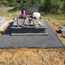 Granitowy nagrobek z czarnym krzyżem i kwiatami, otoczony szarą kostką brukową na piaszczystym podłożu, w tle drzewa.