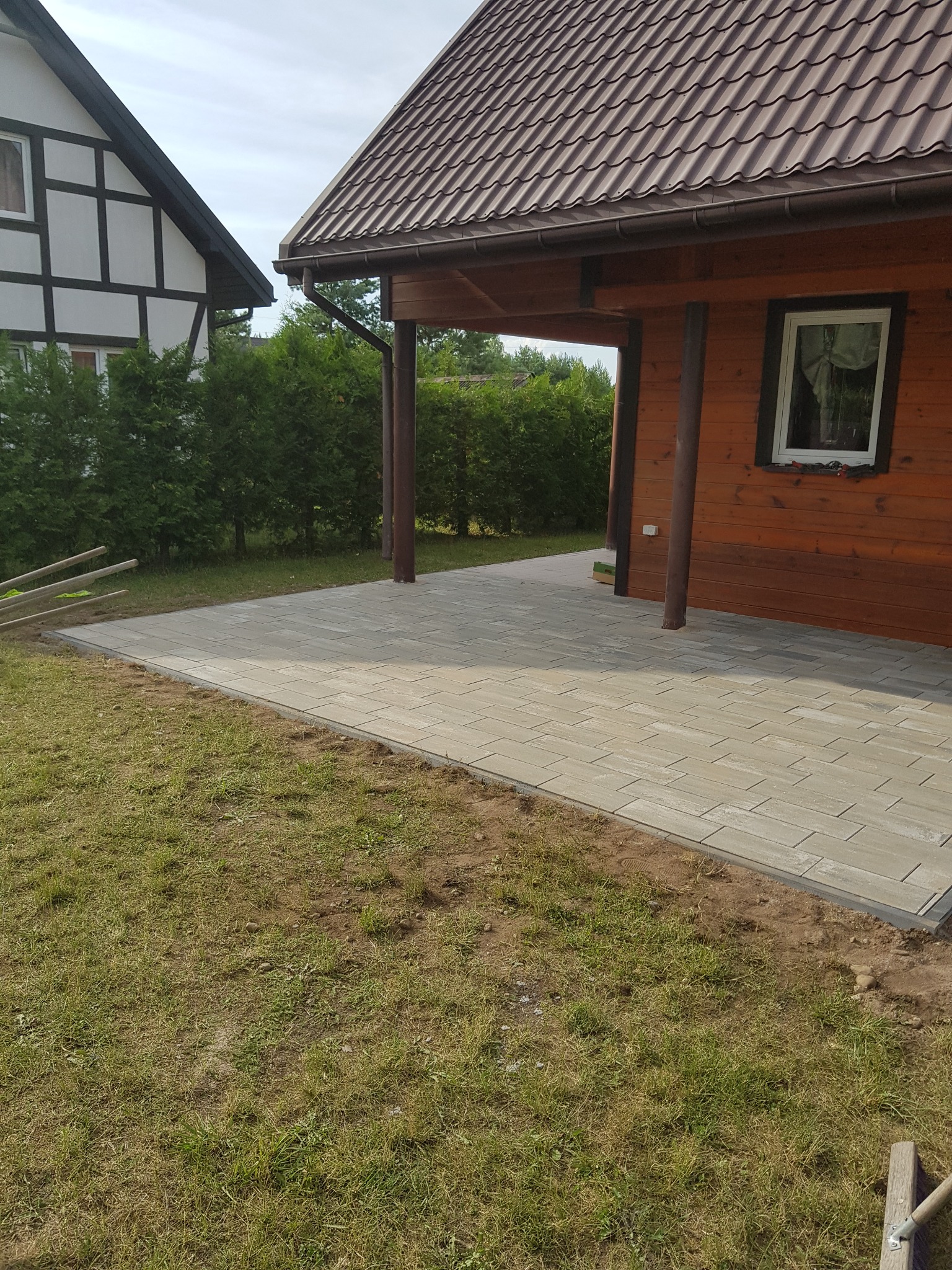 Jasnoszara kostka brukowa ułożona jako taras przy drewnianym domu z brązowym dachem, widoczne narzędzia i trawnik w tle.