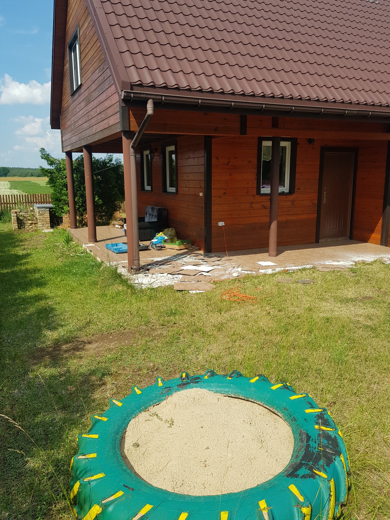 Układanie kostki brukowej na tarasie drewnianego domu; widoczny fragment tarasu z ułożoną kostką, porozrzucane fragmenty kostki oraz piaskownica z opony w trawie.