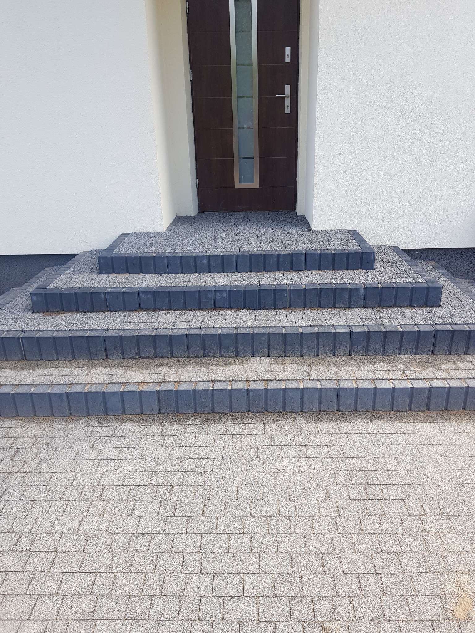 Schody wejściowe wyłożone jasnoszarą kostką brukową z ciemnoszarym obramowaniem, prowadzące do brązowych drzwi wejściowych w białym budynku.