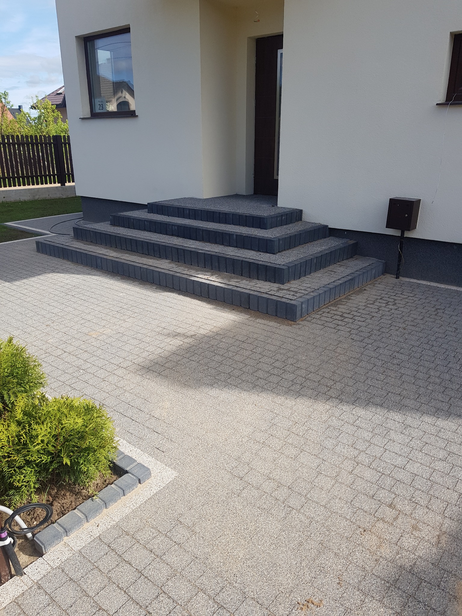 Nowoczesne schody wejściowe i podjazd z szarej kostki brukowej, obrzeża z ciemnej kostki, mały krzew iglasty przy podjeździe.