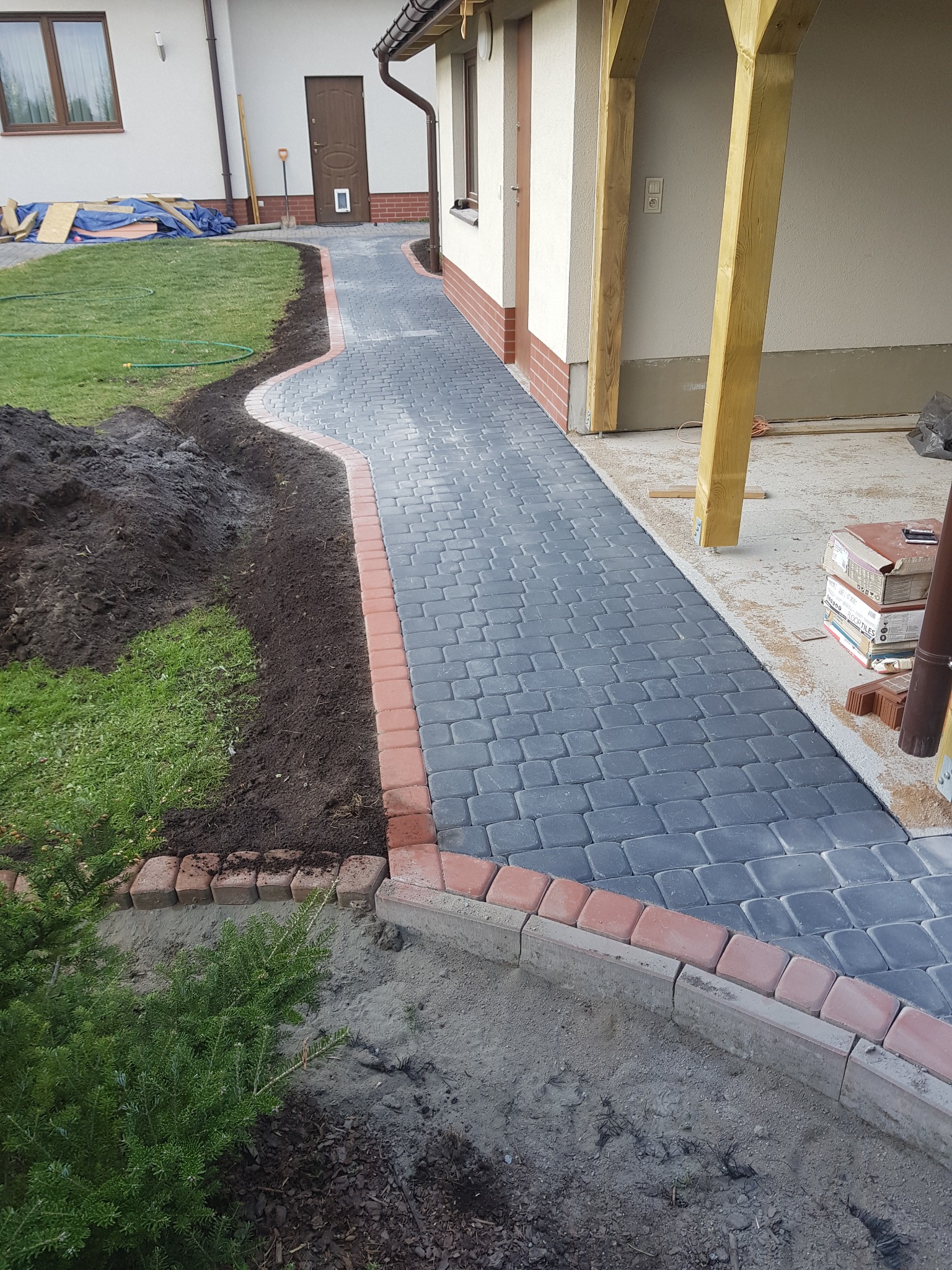 Ścieżka z szarej kostki brukowej z czerwoną obwódką, prowadząca od trawnika do budynku z drewnianą konstrukcją werandy, widoczne prace ziemne po jednej stronie ścieżki.