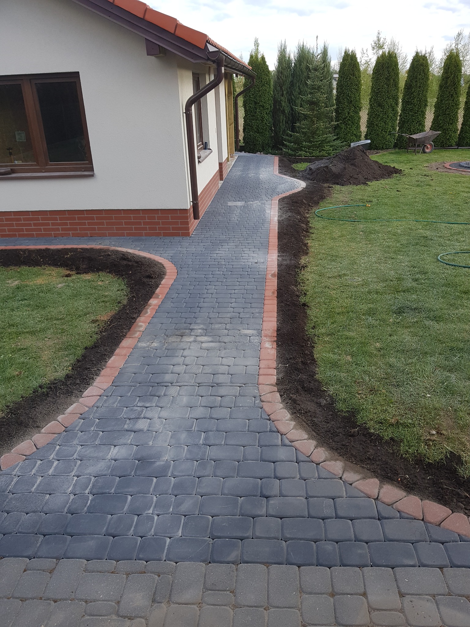 Ułożona ścieżka z kostki brukowej w kolorze grafitowym z czerwoną obwódką prowadząca wzdłuż domu z ceglaną podmurówką, widoczny trawnik z przygotowanym miejscem pod nasadzenia i sterta ziemi.
