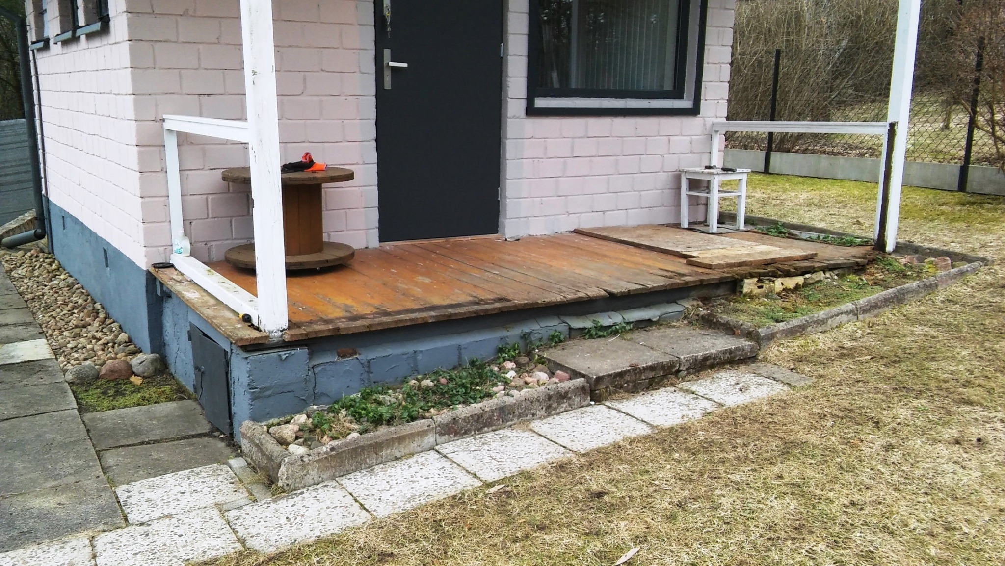 Nierówna drewniana weranda z prowizorycznym stołem z bębna kablowego, schodami z betonowych płyt i obrzeżem rabaty z kamienia.