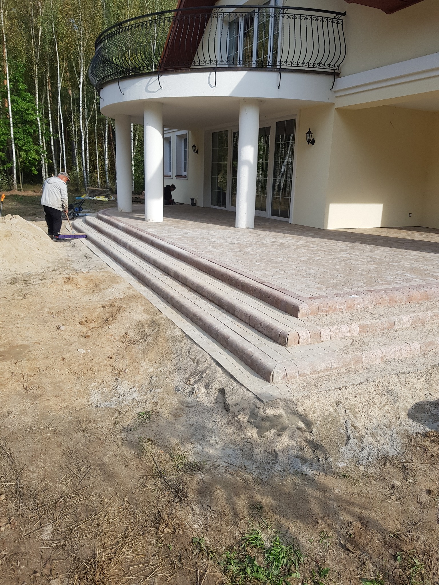 Pracownik zamiata piasek z nowo ułożonych schodów i tarasu z kostki brukowej przed domem z kolumnami i balkonem.