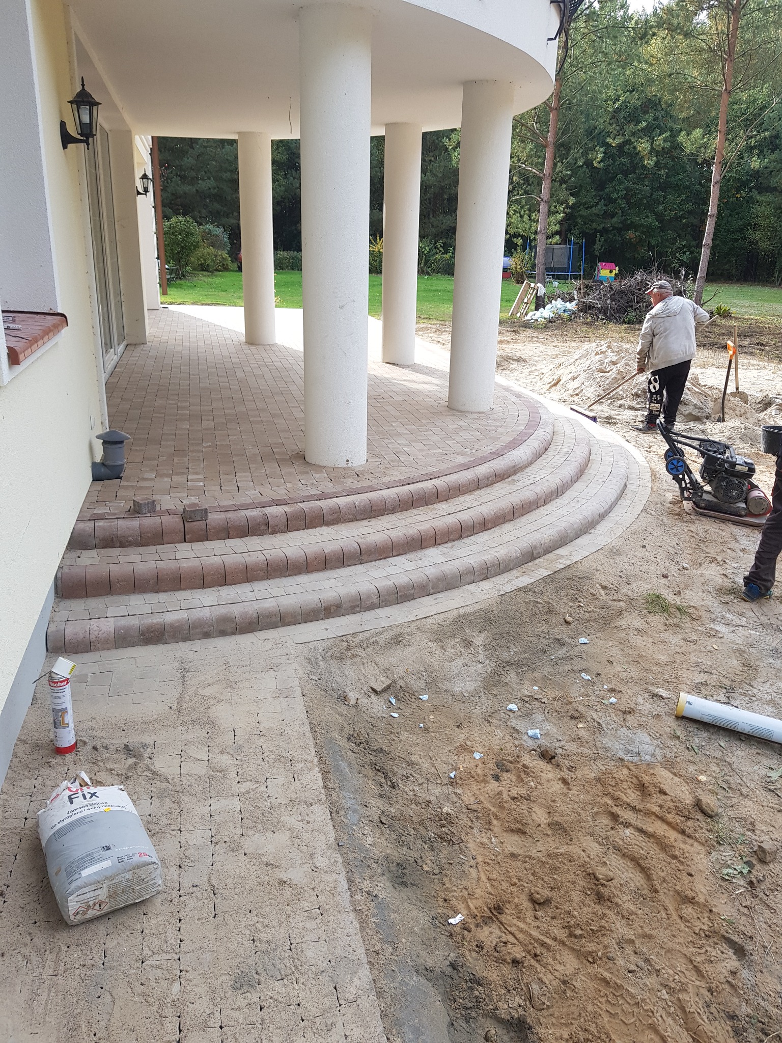 Budowa schodów wejściowych z kostki brukowej przed domem z kolumnami, w tle pracownik ubija ziemię zagęszczarką.