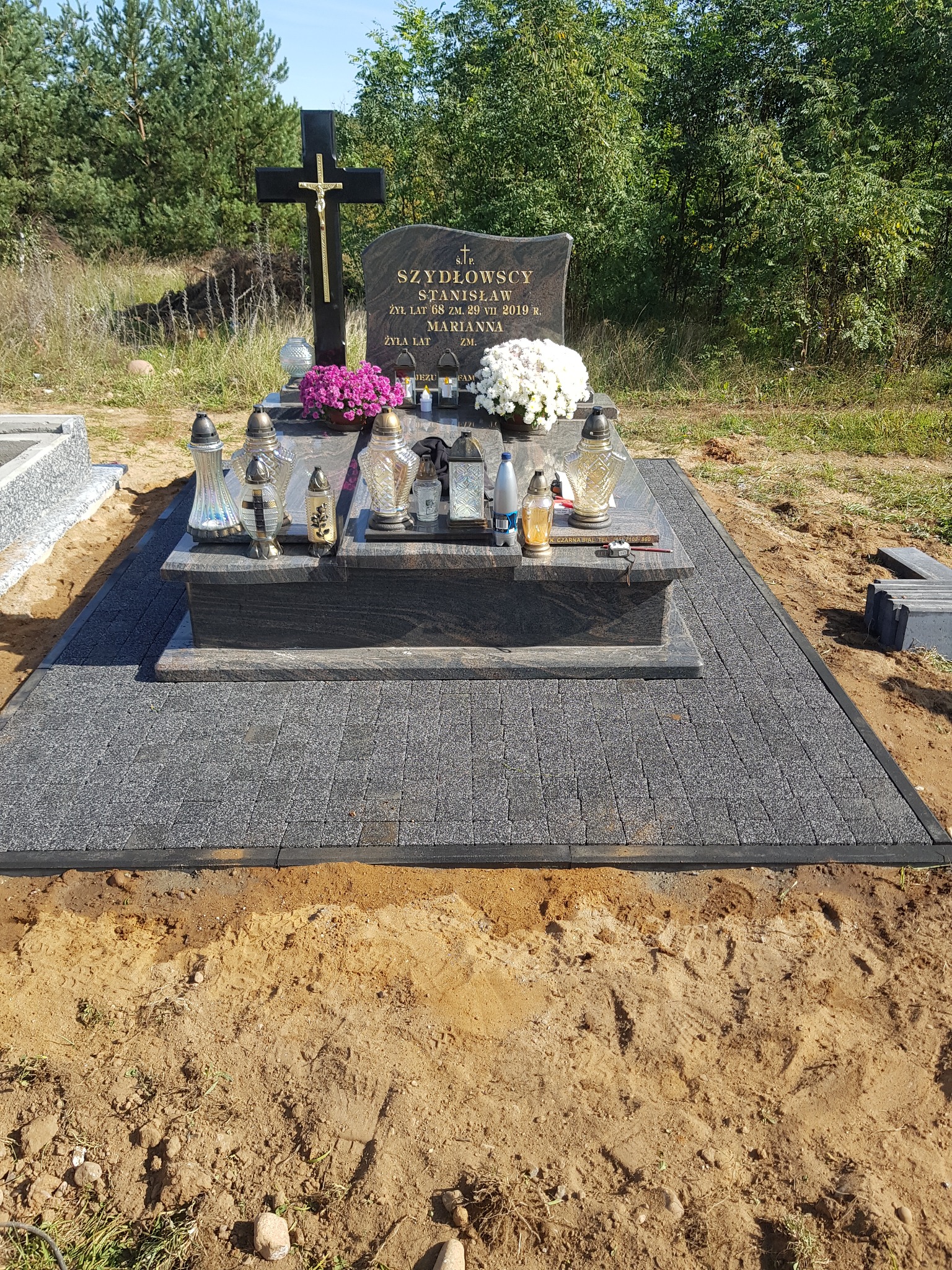 Granitowy nagrobek z czarnym krzyżem i kwiatami, otoczony szarą kostką brukową na piaszczystym podłożu, w tle drzewa.