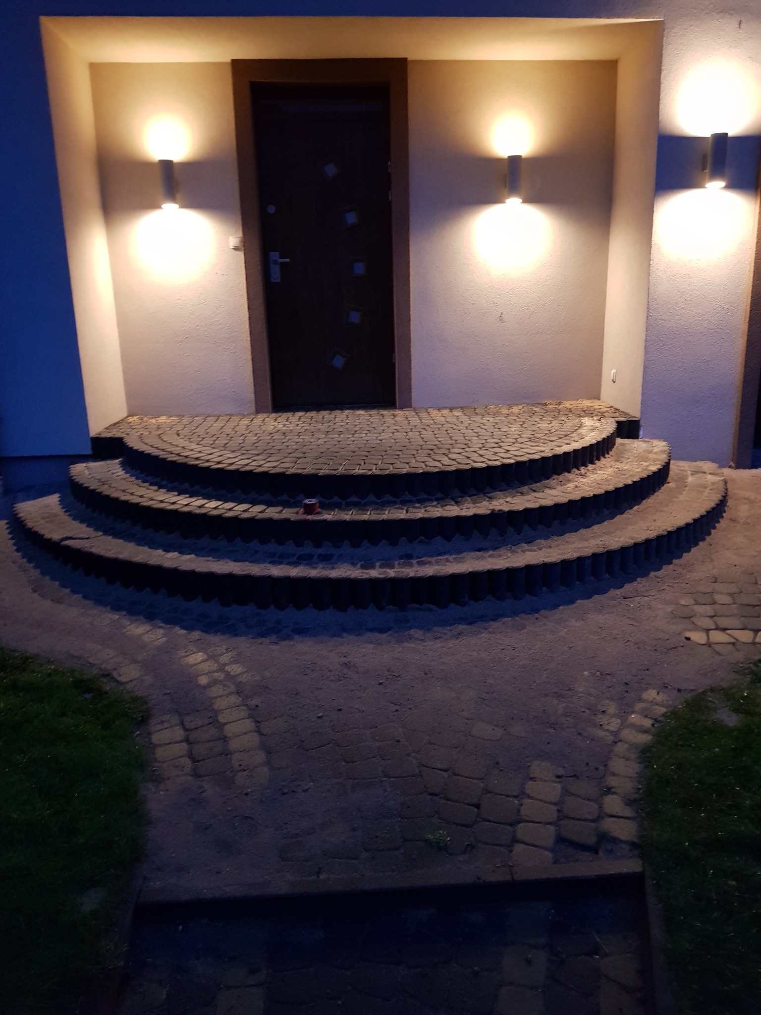 Półokrągłe schody wejściowe z kostki brukowej, oświetlone kinkietami, prowadzące do ciemnych drzwi wejściowych.