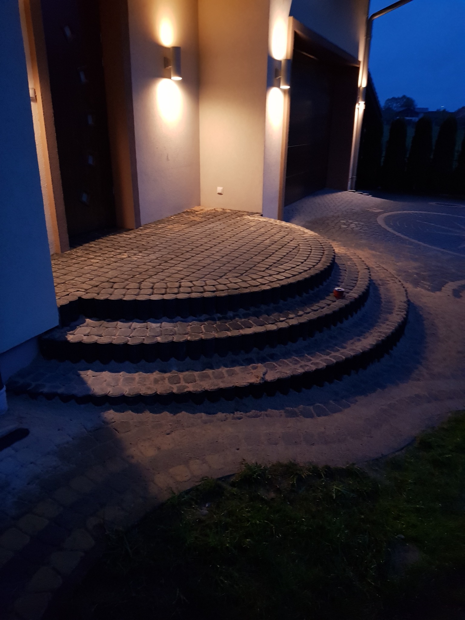 Półokrągłe schody z kostki brukowej prowadzące do wejścia domu, oświetlone ciepłym światłem lamp ściennych, widok na podjazd i ciemne niebo wieczorem.