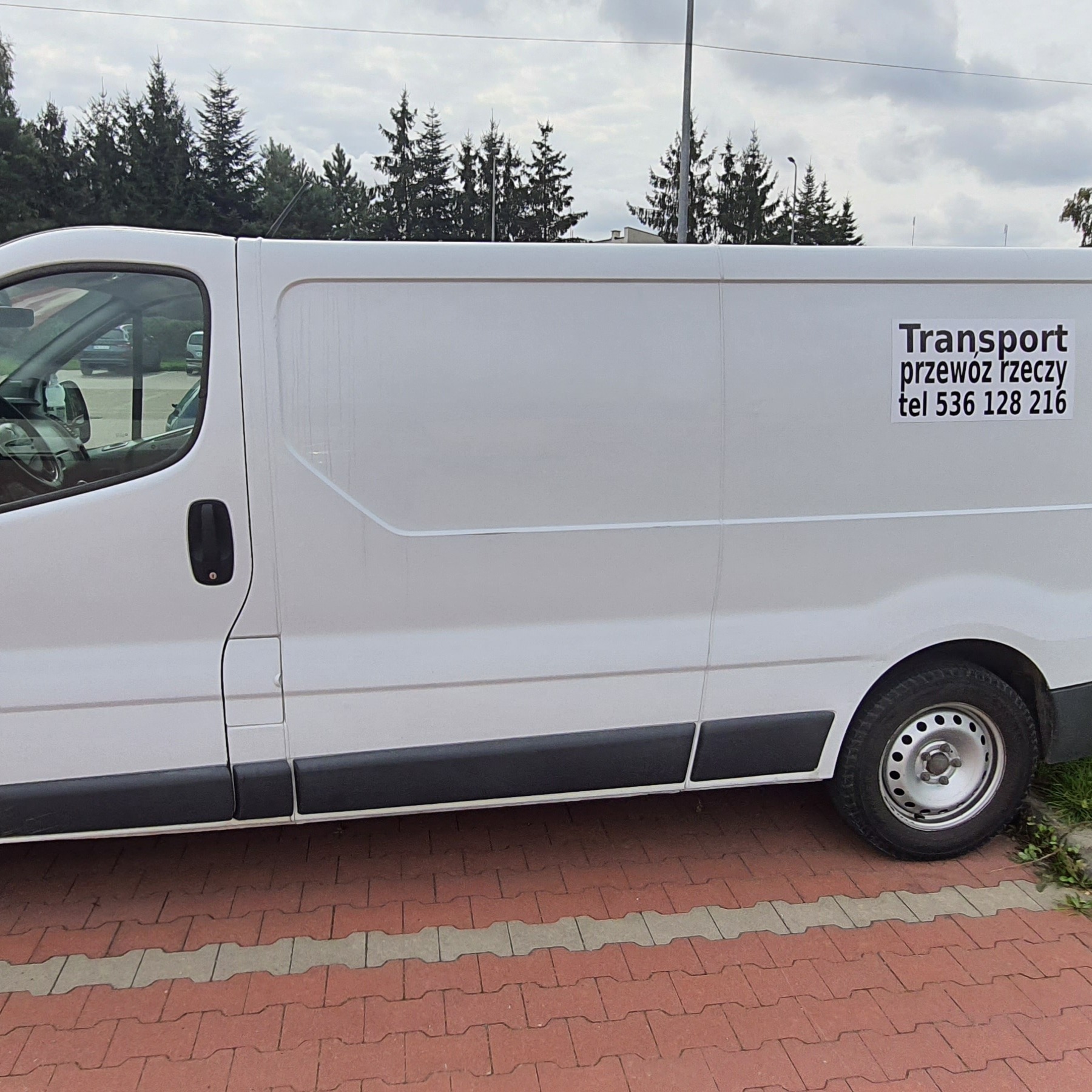 Biały samochód dostawczy z napisem 'Transport przewóz rzeczy tel 536 128 216' na tle drzew iglastych i pochmurnego nieba.