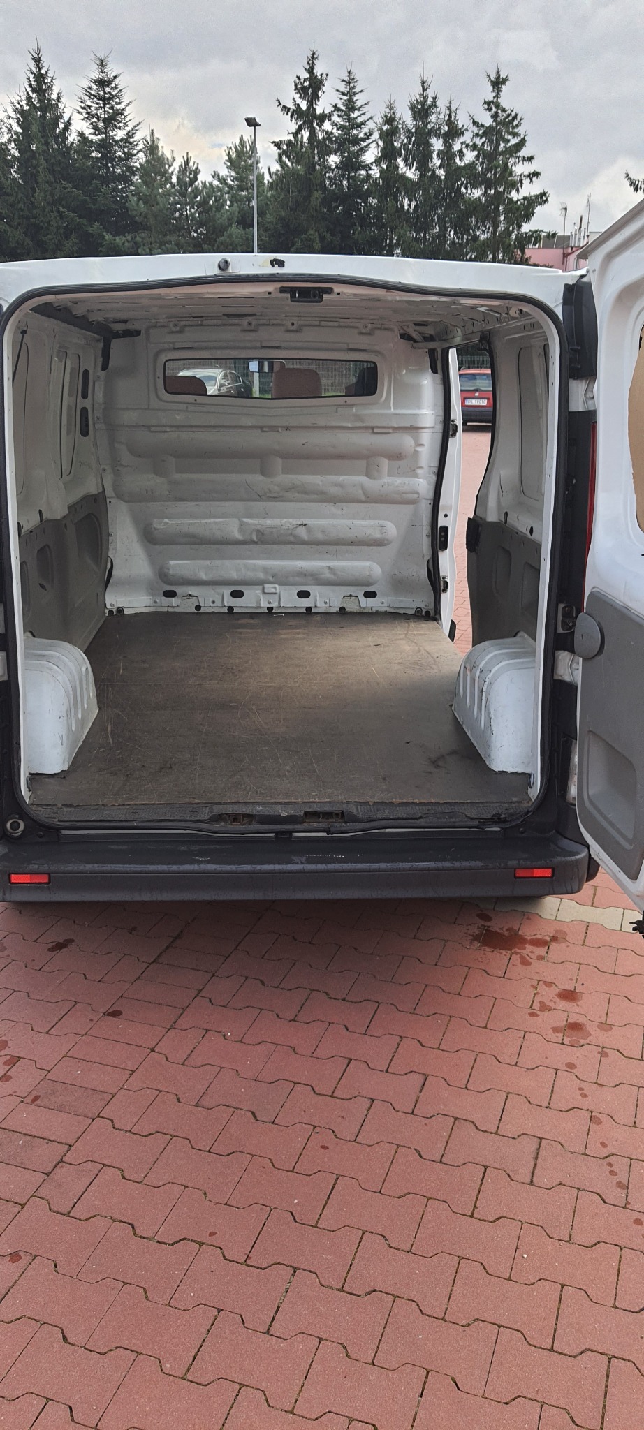 Pusta przestrzeń ładunkowa białego furgonetki z otwartymi tylnymi drzwiami, widok z tyłu, na tle drzew i kostki brukowej.