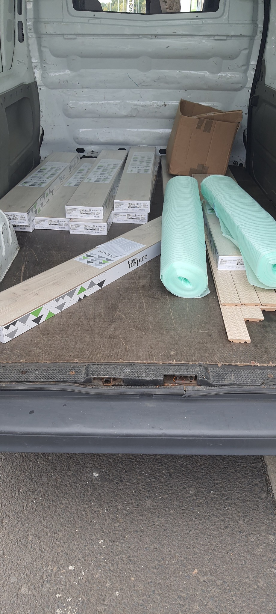 Wnętrze furgonetki załadowane pakietami paneli podłogowych marki Home Inspire, rolkami pianki izolacyjnej i kartonowym pudłem, przygotowane do transportu.