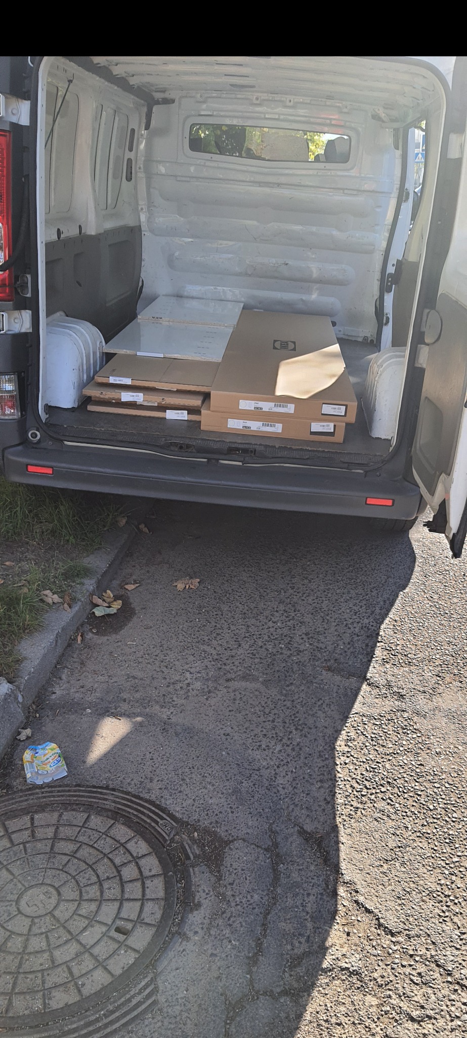 Wnętrze białego busa z otwartymi tylnymi drzwiami, załadowane płaskimi, kartonowymi paczkami z widocznymi etykietami, prawdopodobnie meble do samodzielnego montażu.