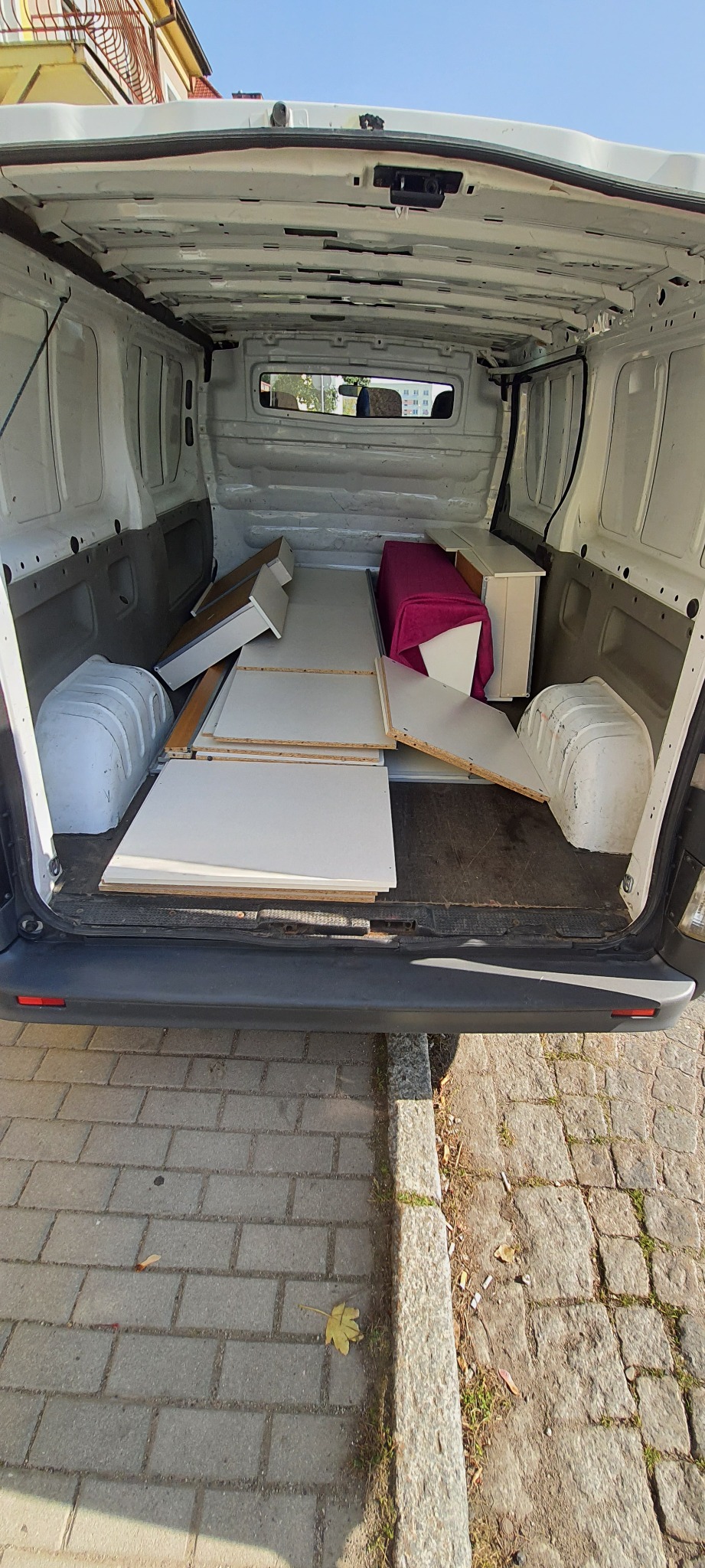 Wnętrze furgonetki załadowane elementami meblowymi, w tym białymi płytami i szafką przykrytą różowym materiałem, widoczne z zewnątrz pojazdu na tle chodnika z kostki.