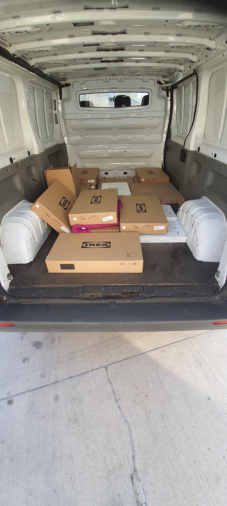 Wnętrze białego furgonetki wypełnione kartonami z logo IKEA, częściowo przykrytymi różowym kocem, sugerujące transport mebli.